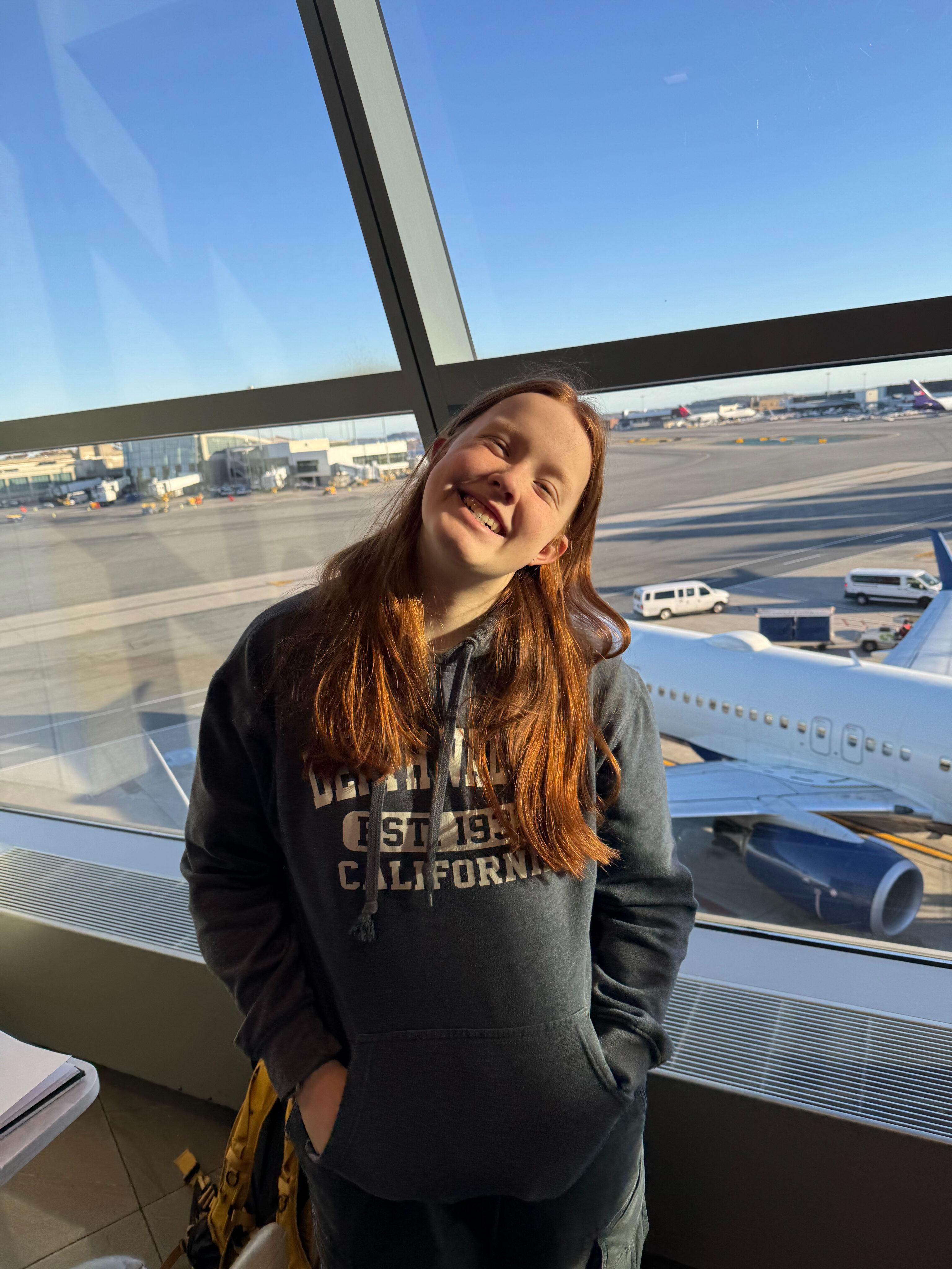 Cameron wearing a Death Valley sweatshirt, standing and smiling poses for a photo in the Delta Sky Club with giant windows and airplanes behind her.