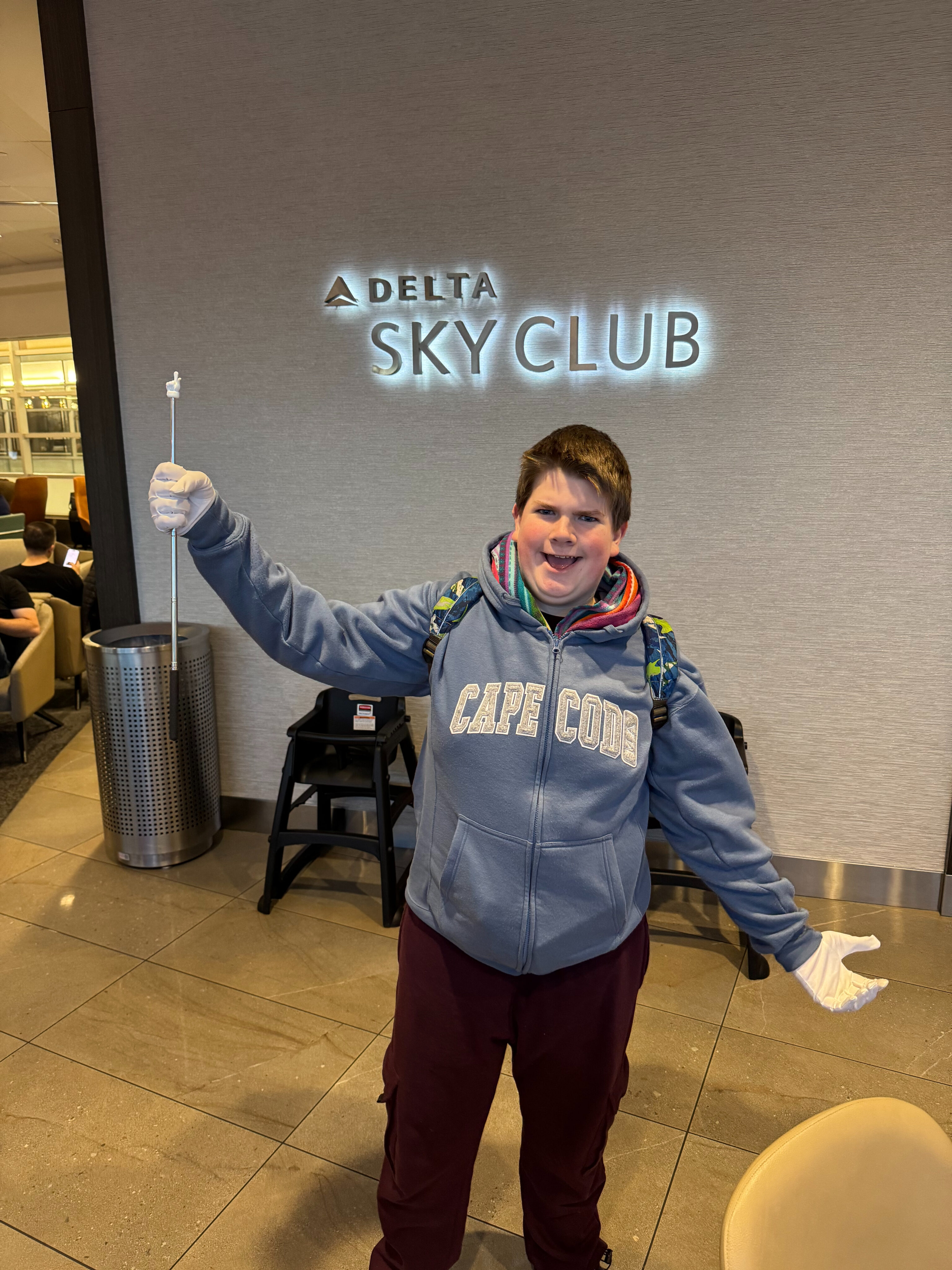 Collin where a Cape Cod Sweatshirt smiles and poses for a photo in front of the Delta Sky Club neon sign. 