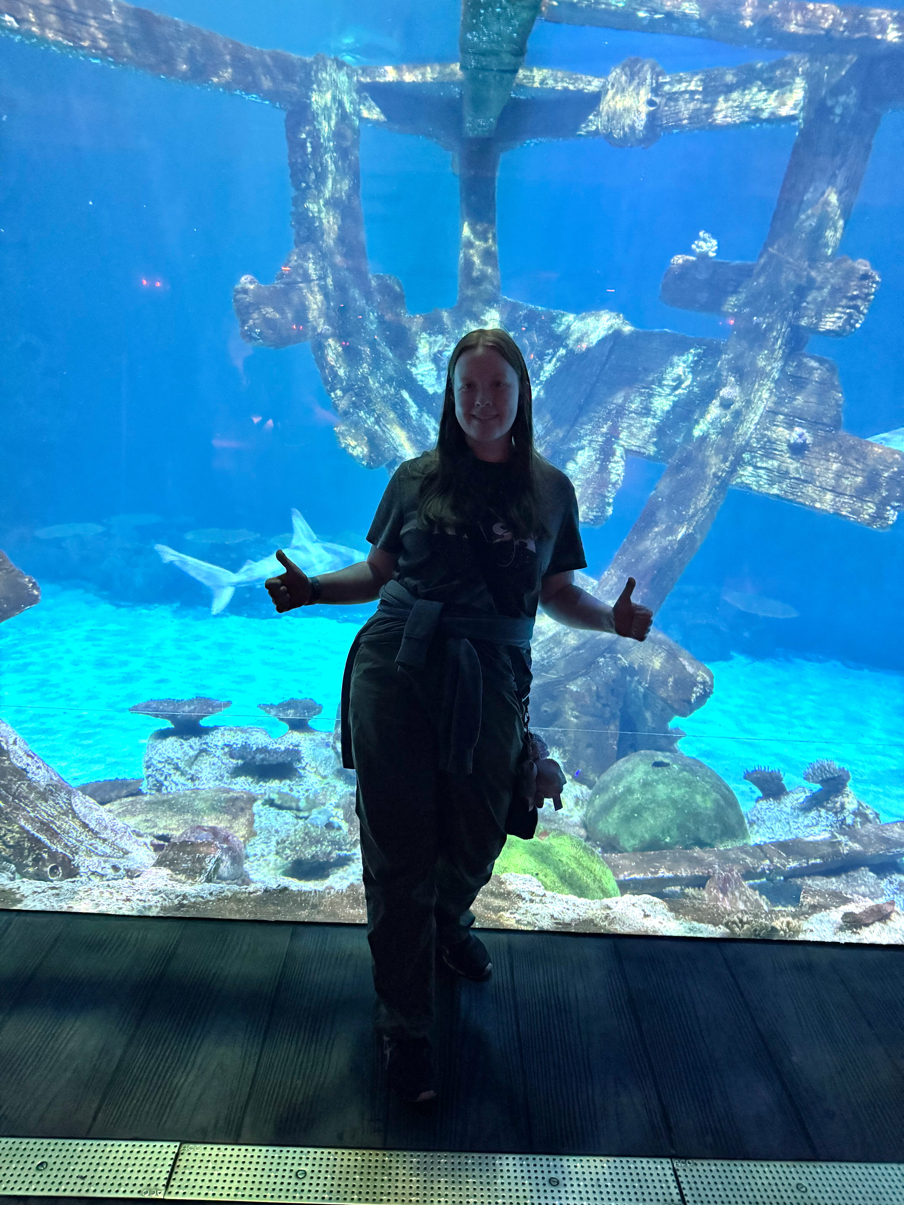 Cameron standing with a smile and giving two thumbs up in front of massive fish tank at the Shark Reef Aquarium.