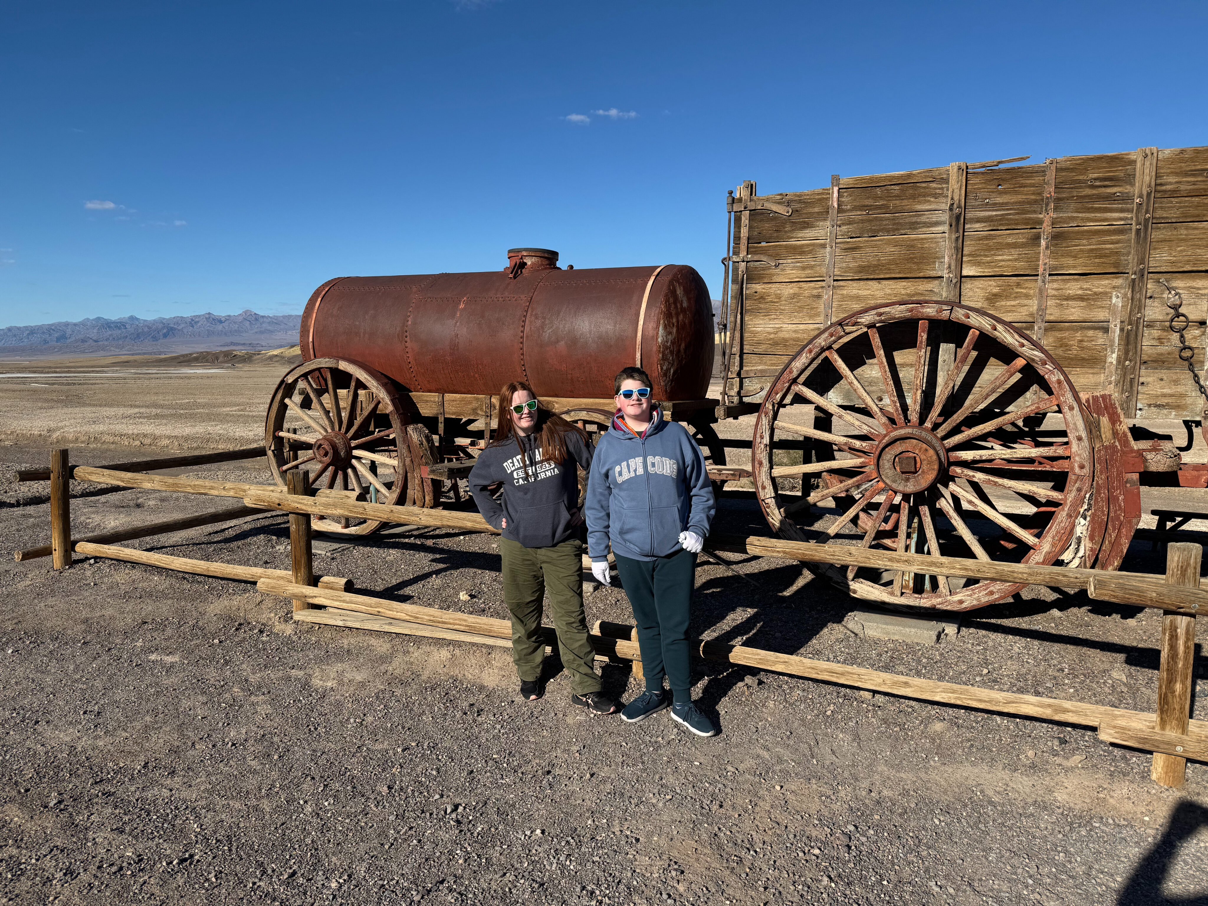 Collin and Cameron wearing sunglasses and standing in front of the twenty mule team wagon at the Harmony Borax Works in Death Valley.