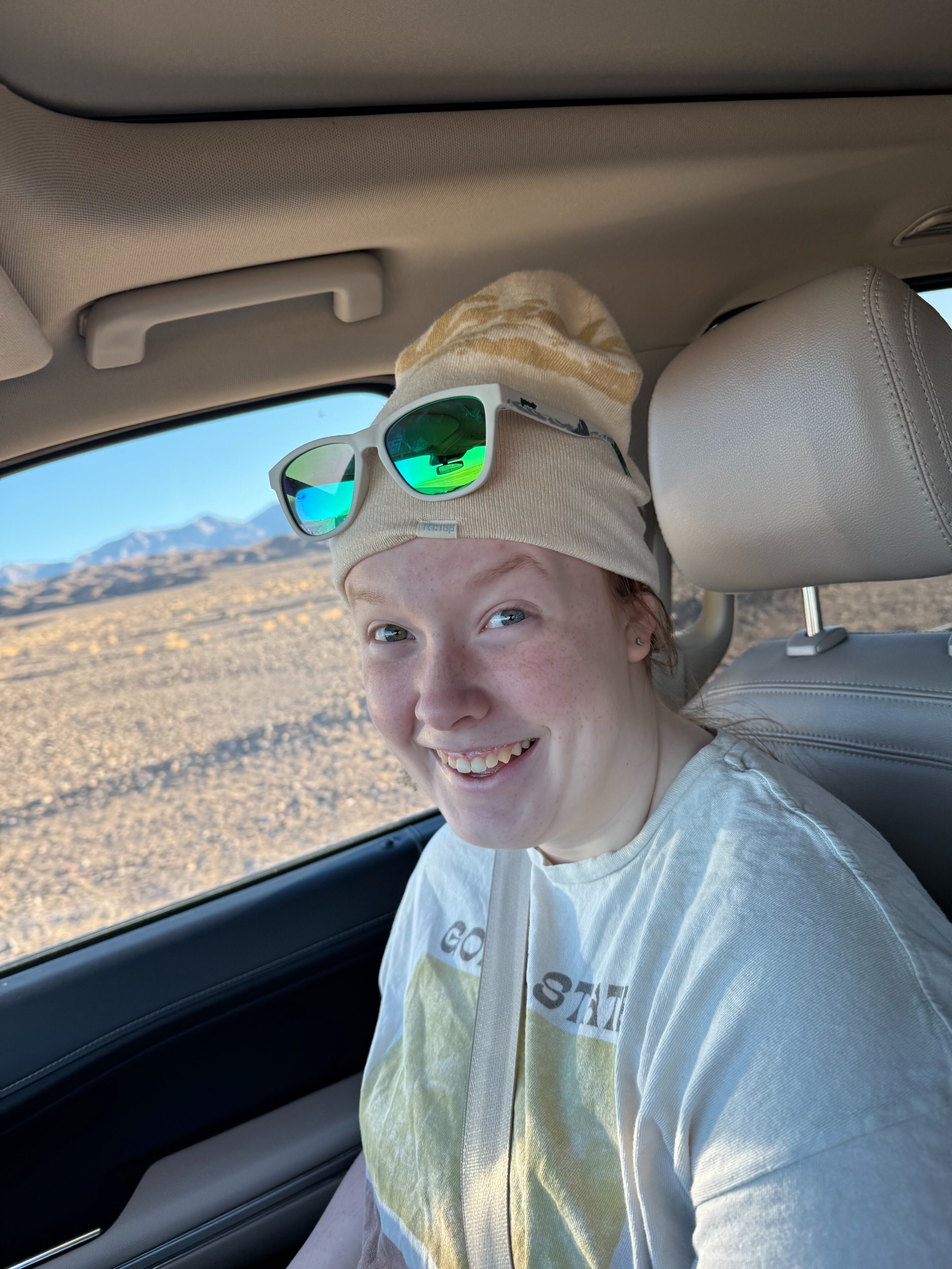 Cameron sitting in the car, with a big smile on her face, winter hat and sunglasses on driving through Death Valley.