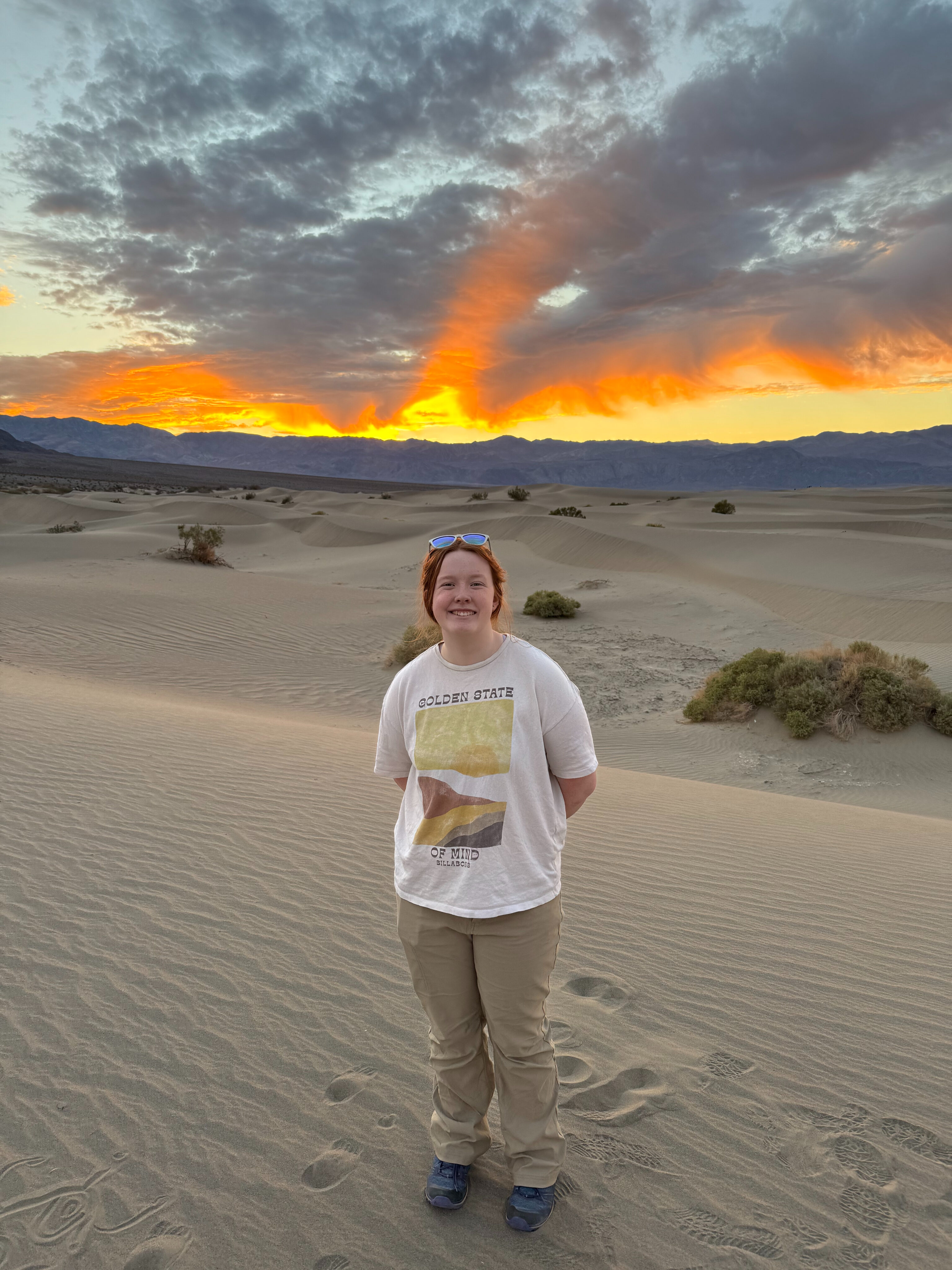 Cameron standing on the sand at the Mesquite Dunes at sunset with clouds and a red sky behind her.