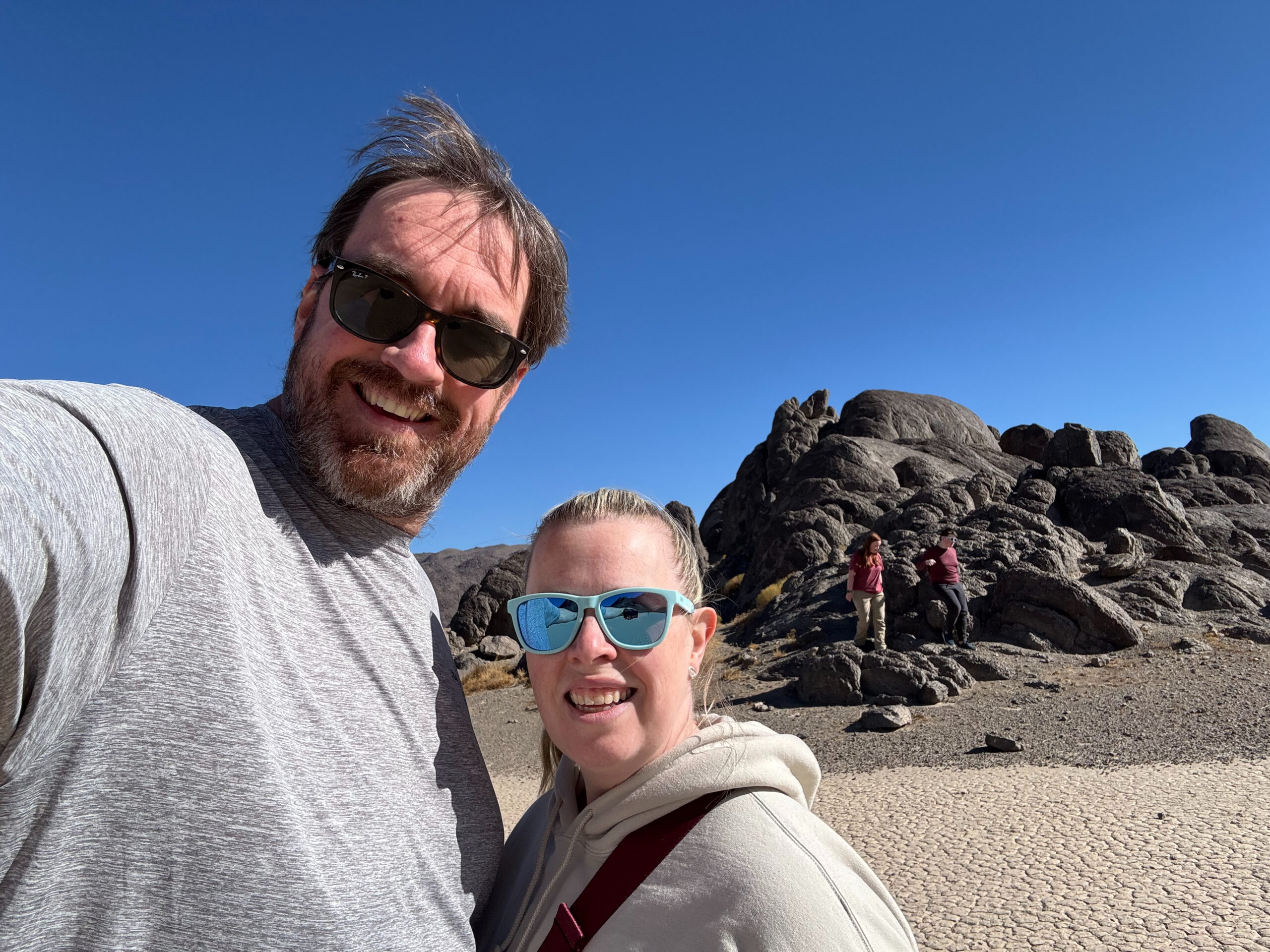 Chris and Cat pose for a photo wearing sunglasses on the Racetrack in Death Valley just behind them is the Grandstand. 