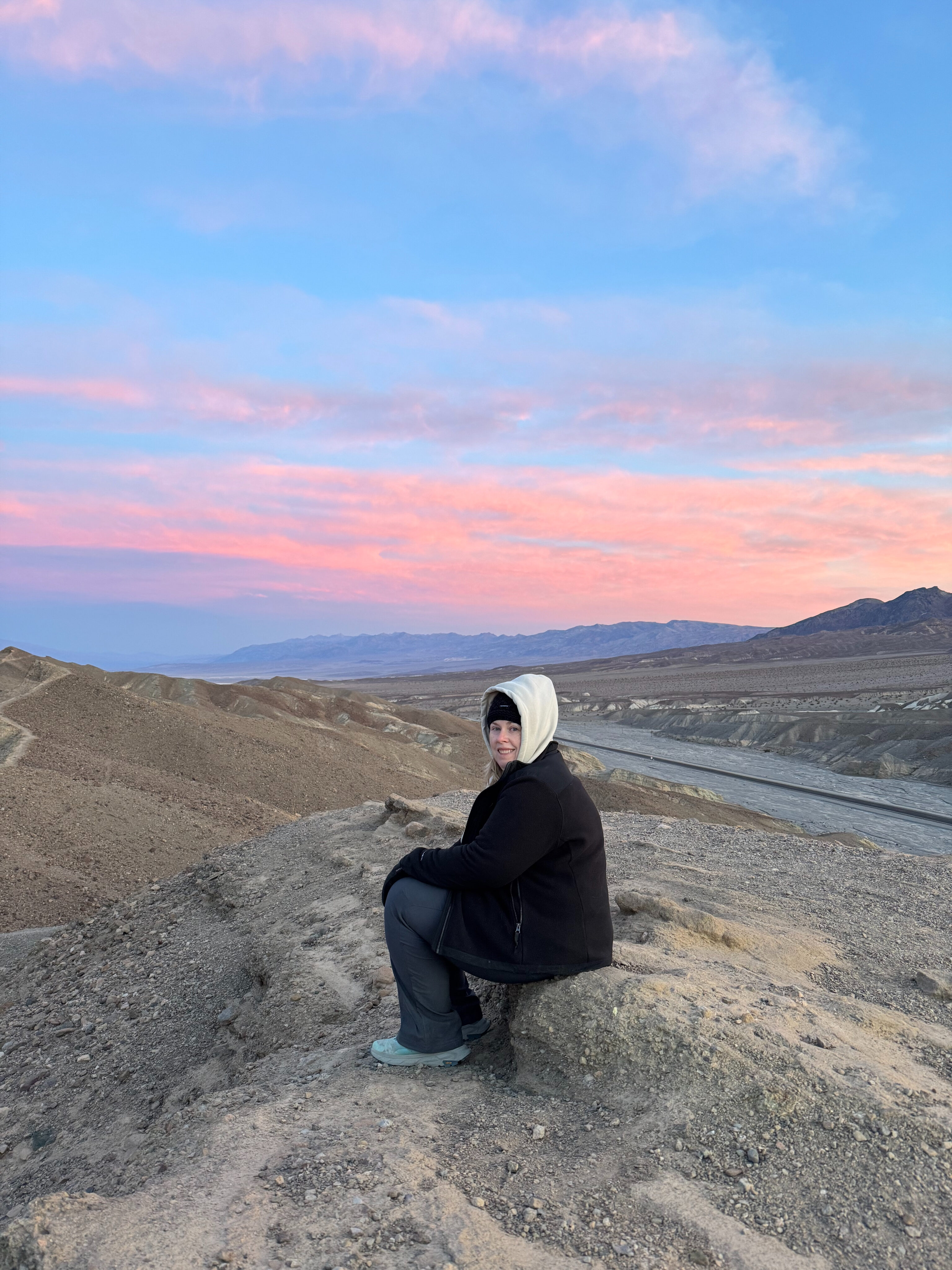 Cat sitting all bundled up in winter gear on top of the badlands at Zabriskie Point in Death valley with a red sunrise sky behind her.