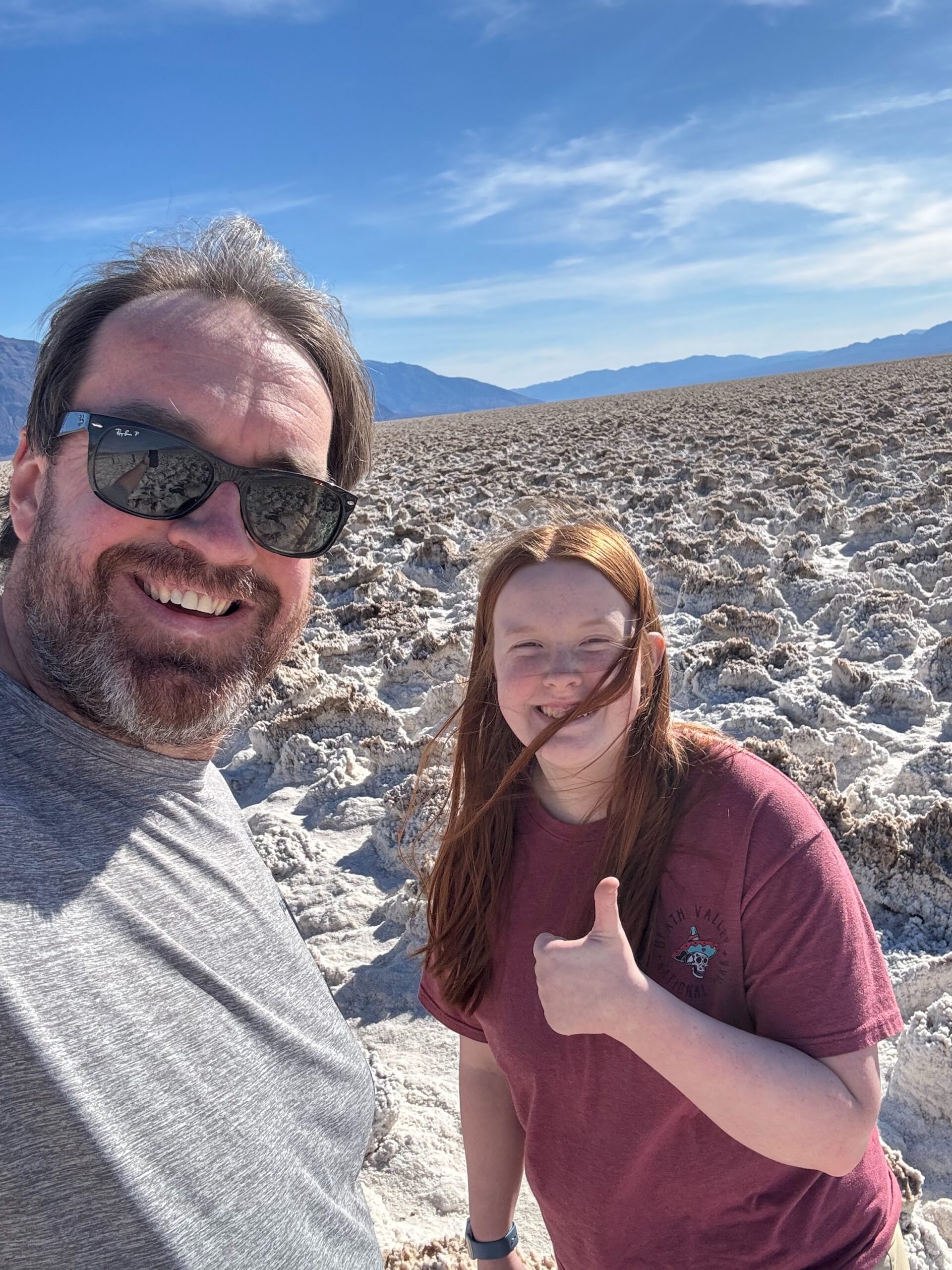 Chris wearing sunglasses and Cami giving a thumbs up standing in the middle of the Devils Golf Course in Death Valley with blue sky and clouds above.