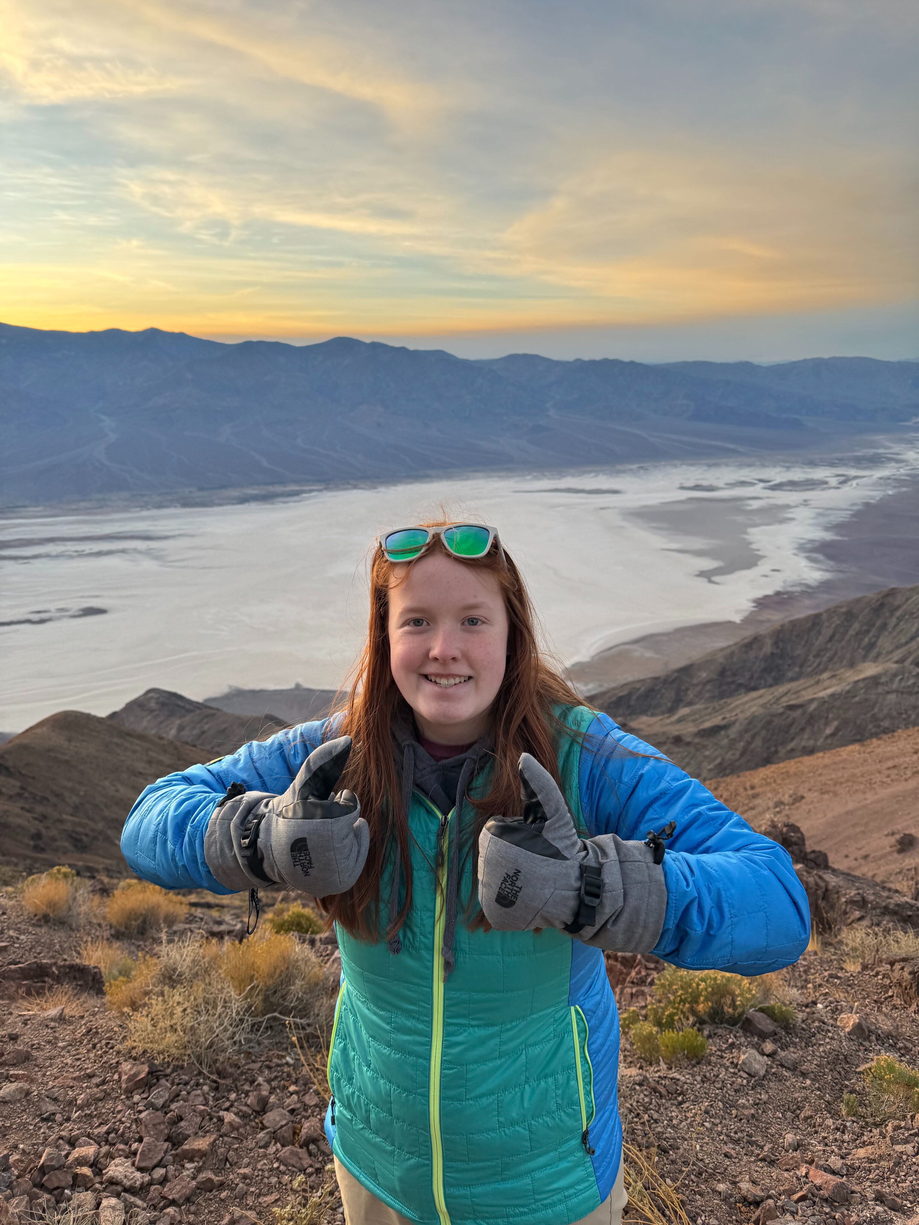 Cami in her winter gear giving two thumbs up with gloves - at Dante's View with a bright sunset sky behind her.