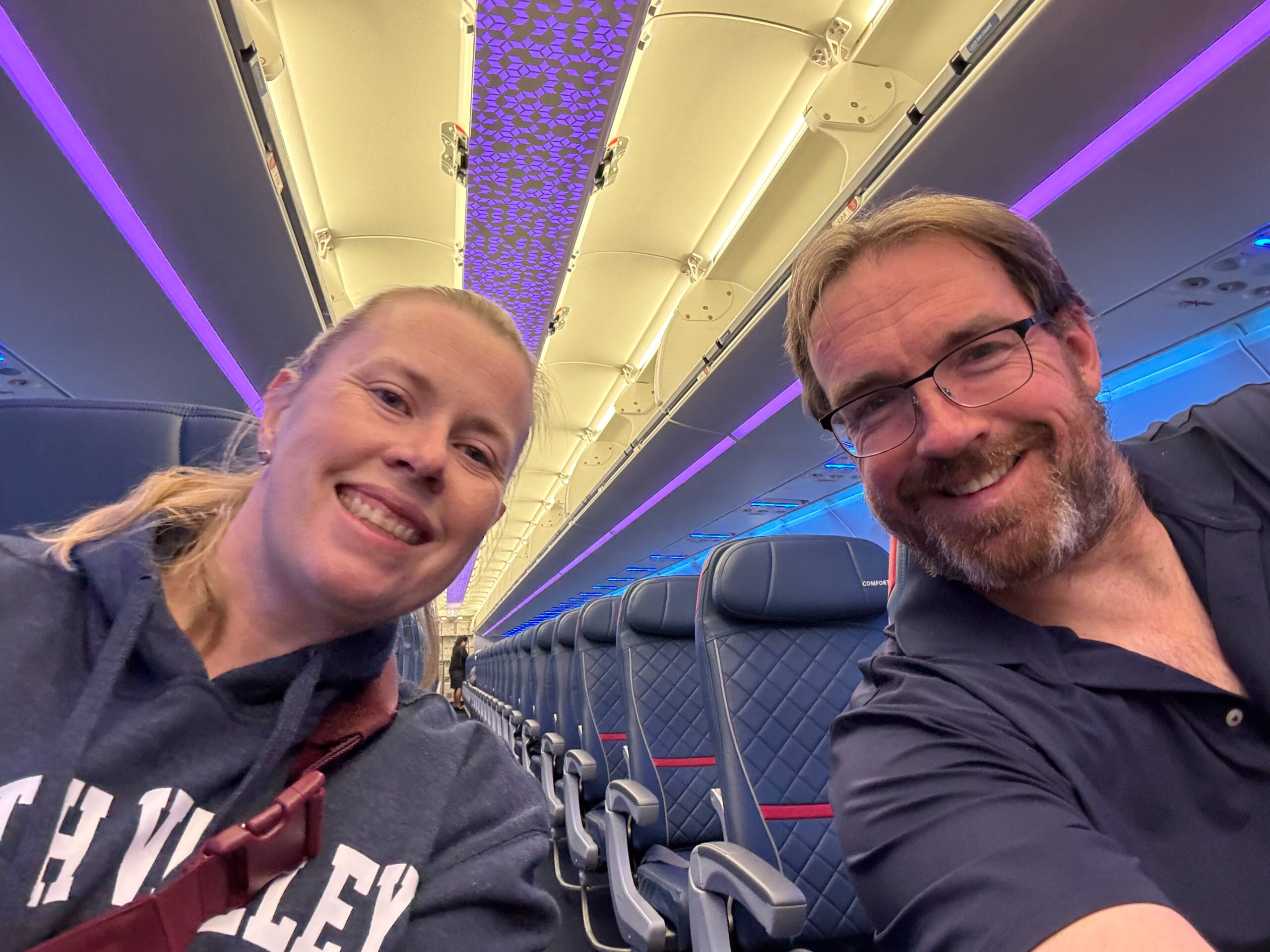 Cat and Chris sitting on different sides of the isle lean in for a photo on the Delta flight home.