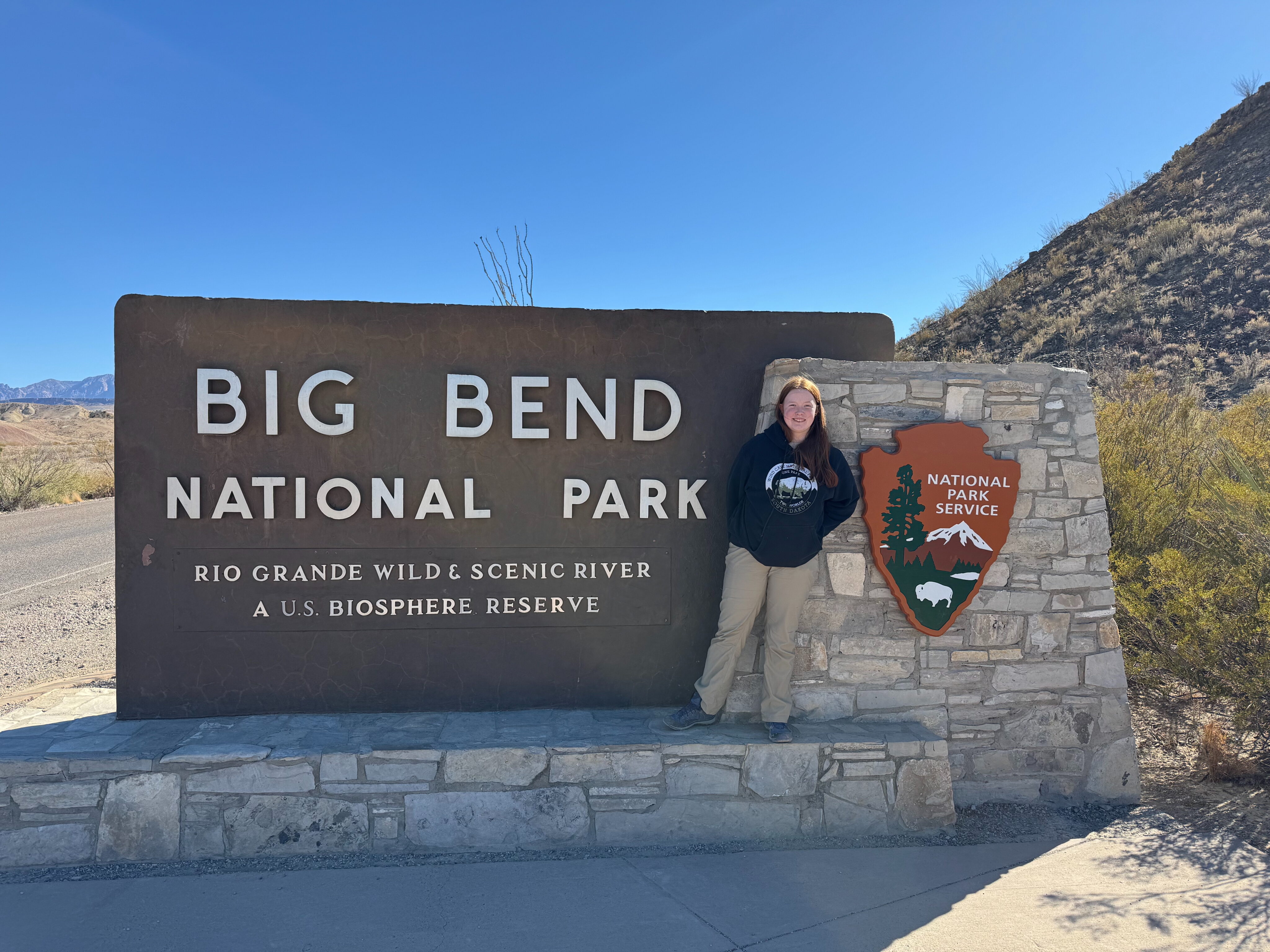 Cameron in hiking pants and a sweatshirt standing next to the Big Bend National Park sign on a blue sky day.