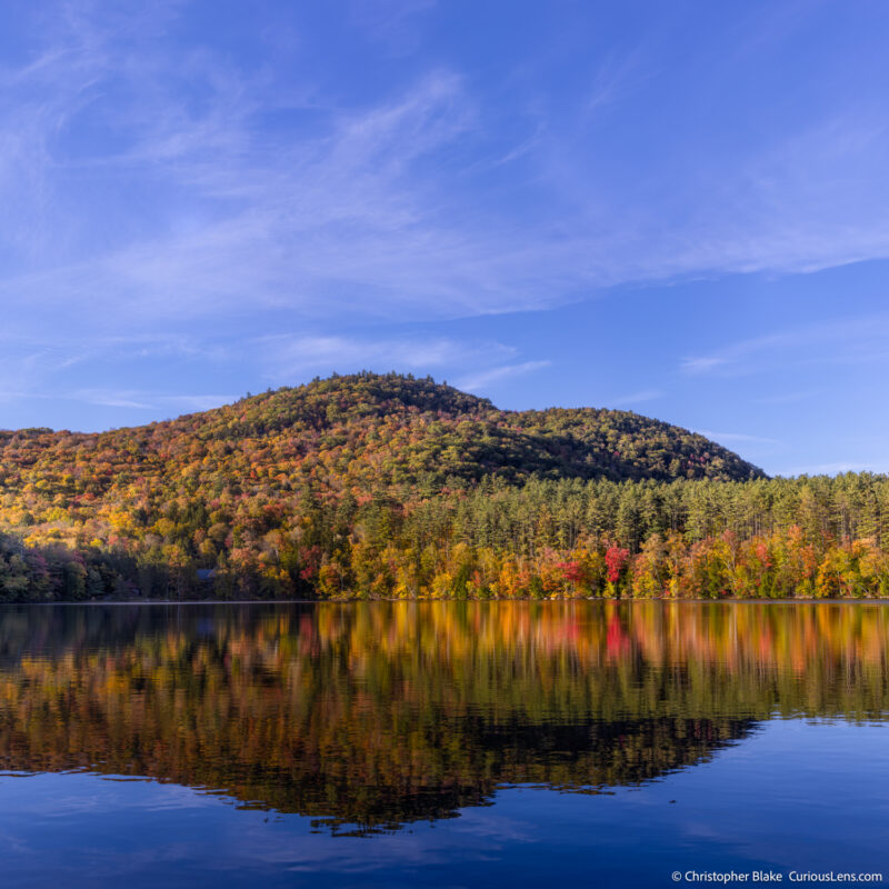 End-of-day view of Mirror Lake in the White Mountains reflecting the vivid autumn colors with soft ripples on the water surface and wispy clouds in the sky.