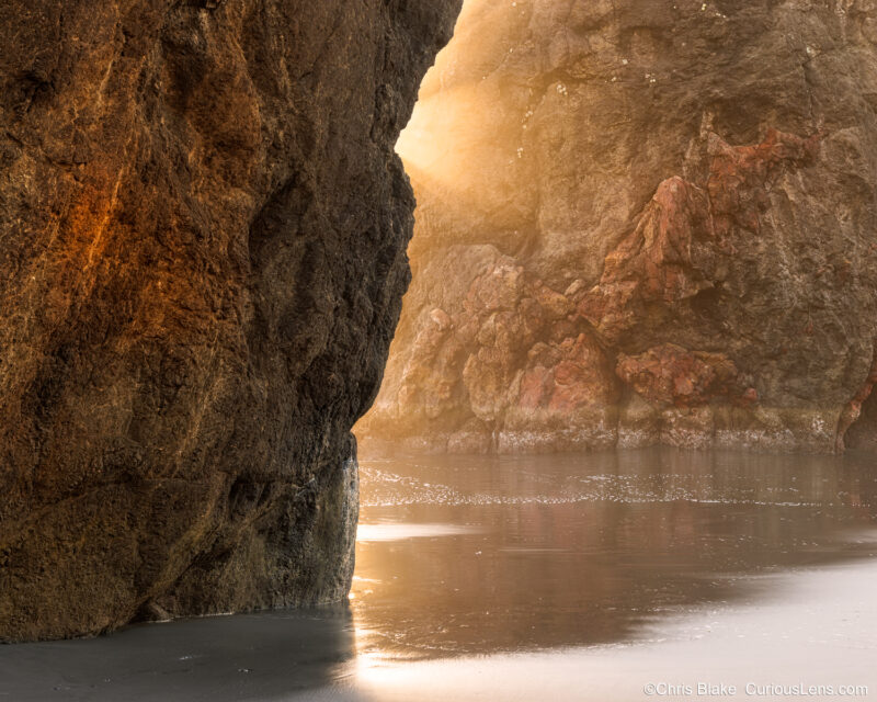 Sunlight beams between sea stacks at Ariya Beach, Oregon, with reflections on wet sand in the foreground.
