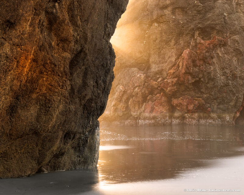 Sunlight beams between sea stacks at Ariya Beach, Oregon, with reflections on wet sand in the foreground.