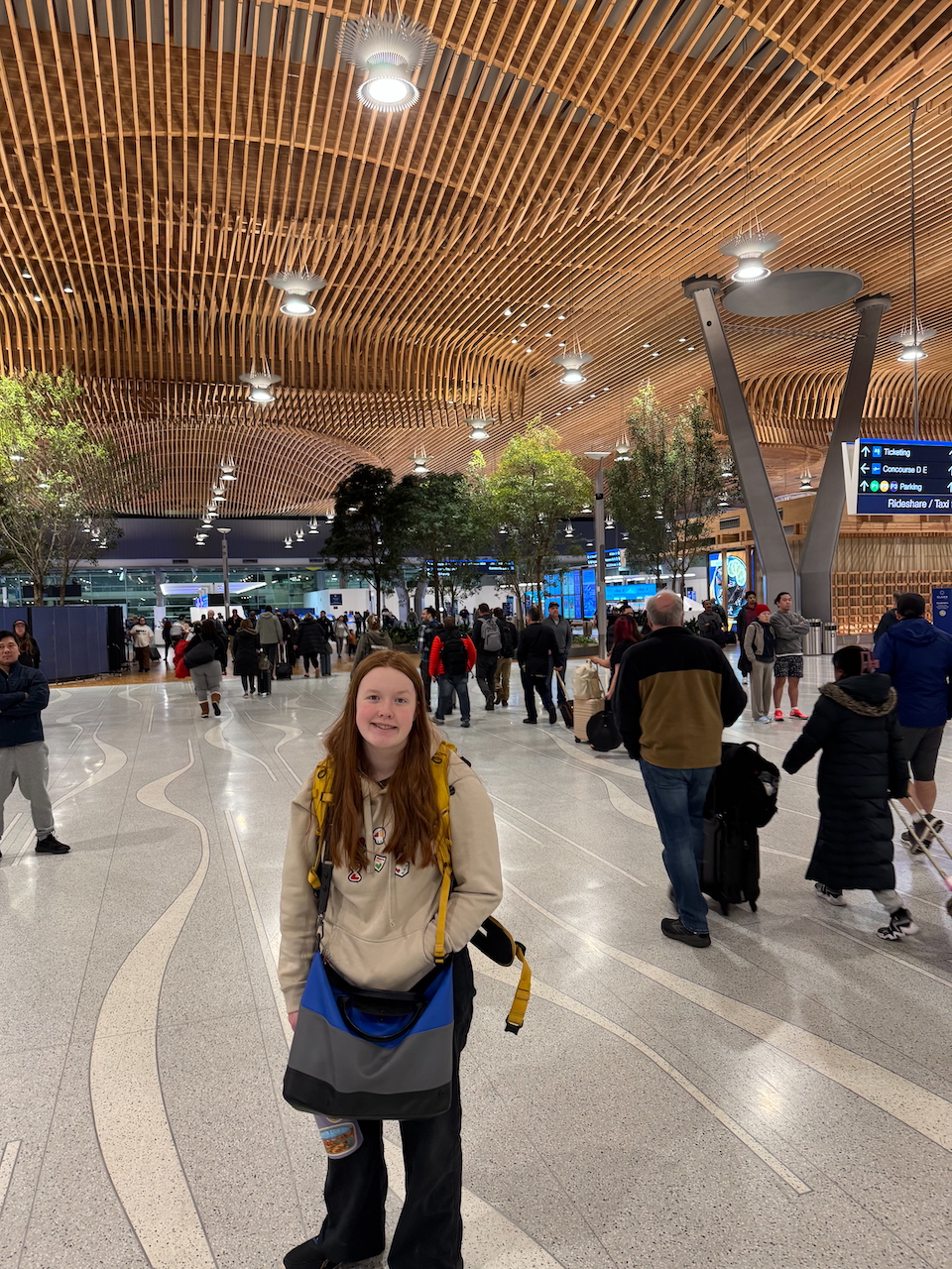 Cameron standing in the middle of the Portland Oregon Airport Terminal. Wearing a hoodie, yellow camera backpack and messenger bag.