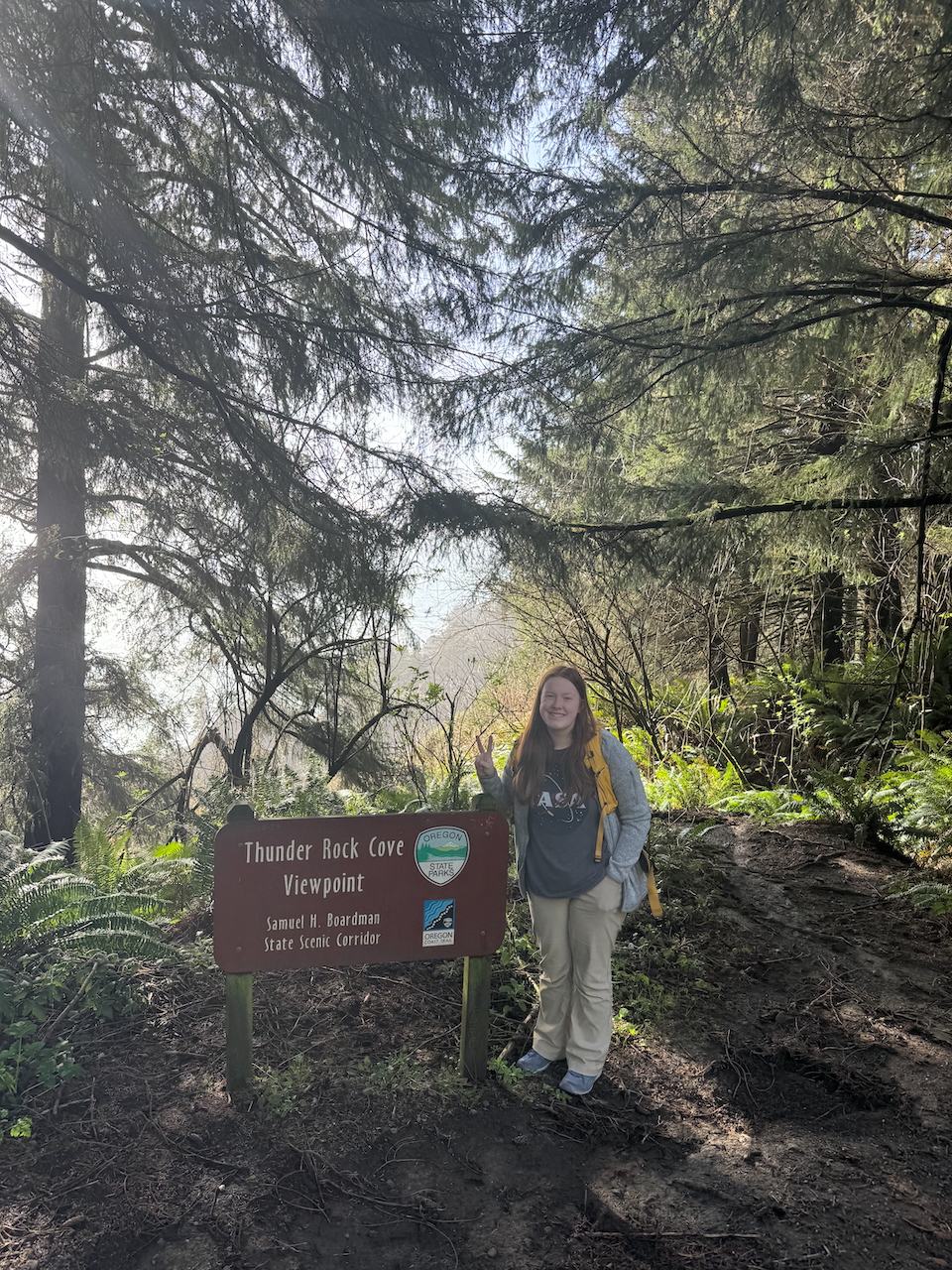 Cameron dressing in hiking clothing, with her camera backpack making a piece sign at the sign post for the Thunder Rock Cove Trail.