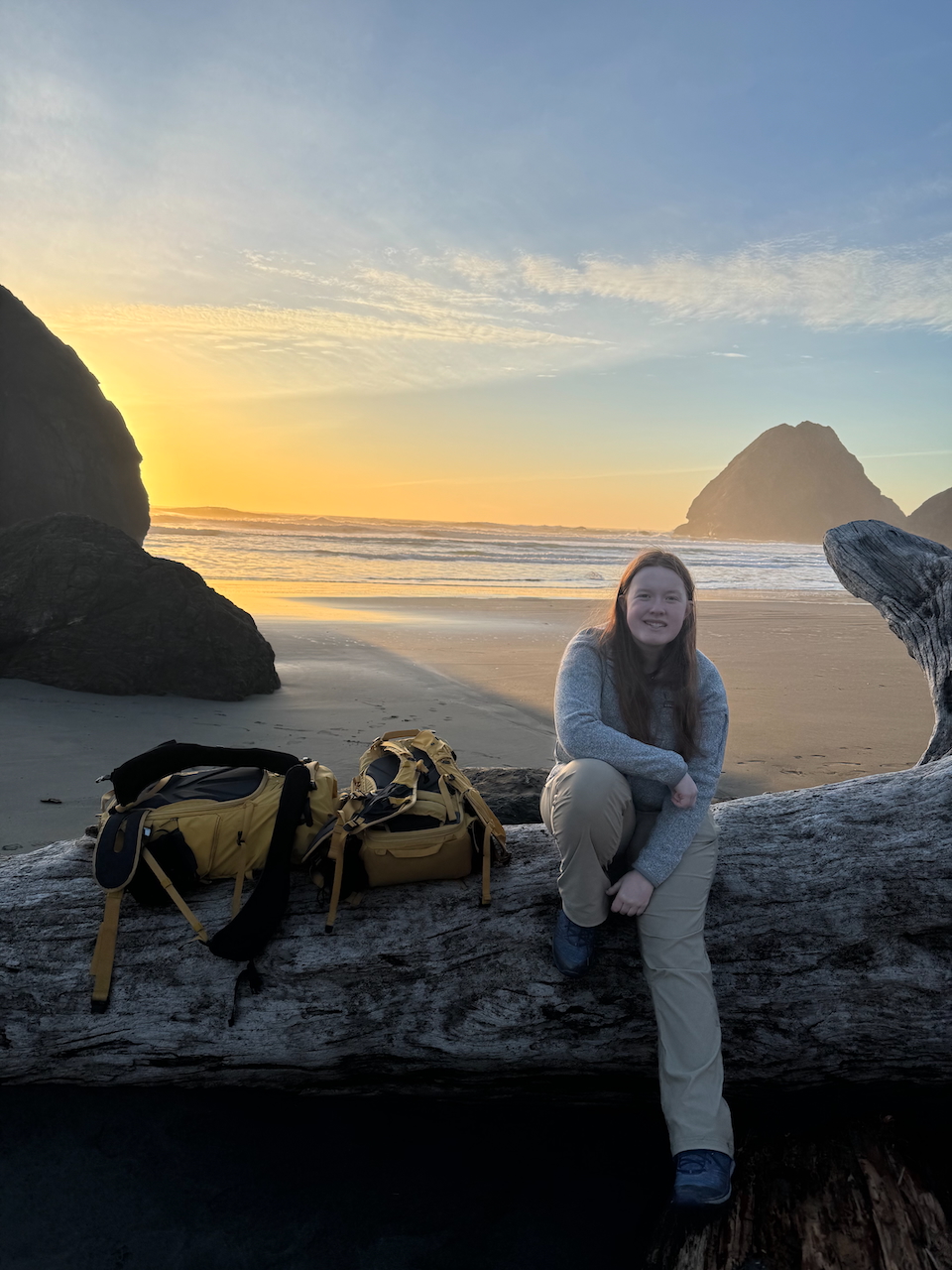 Cameron sitting on a log on Ariya's Beach at sunset with sea stacks and golden light rays behind her.