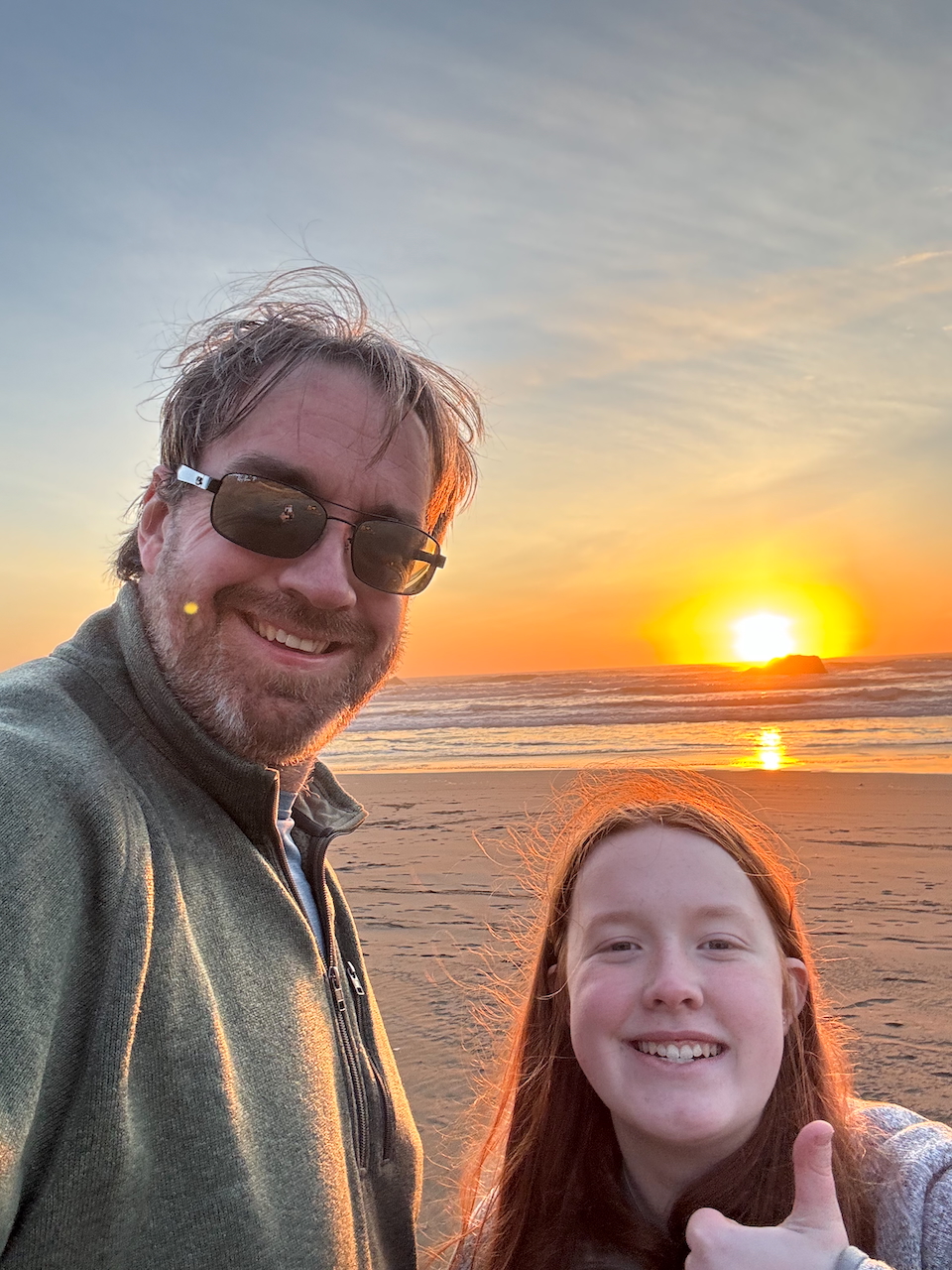 Cameron giving a thumbs up and a big smile along with myself with the waves crashing behind us and then sun setting just above the horizon at Ariya's Beach Oregon.