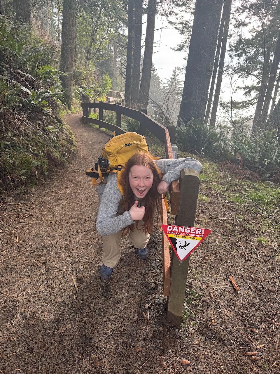 Cameron with a camera bag, on a hiking trail, next to a railing and a Danger of falling sign with a big smile and giving a thumbs up.