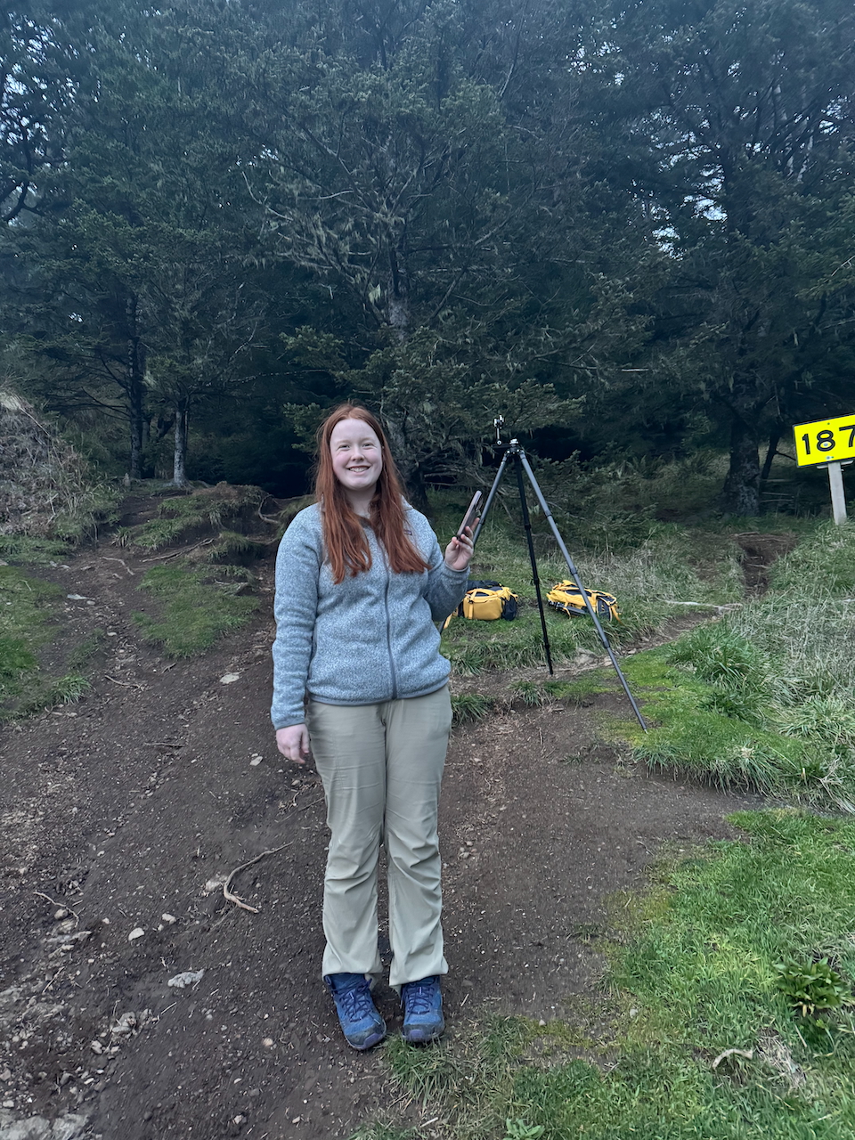 Cameron standing on a hill in her hiking cloths at Secret Beach with a big smile and our camera gear and tripod in the background.