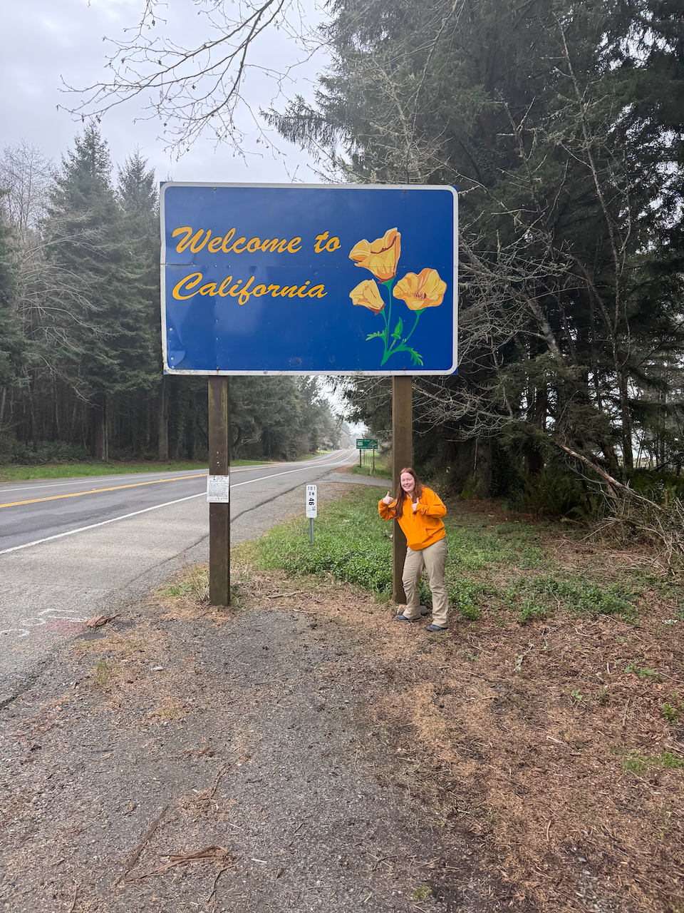 Cameron standing on the side of the 101 and the Welcome to California Sign.
