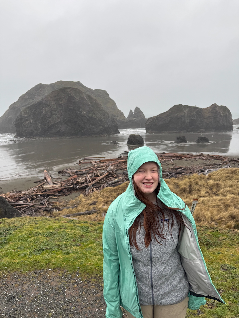 Cameron in a raincoat standing on the grass just above the drift wood and sea stacks on Ariya's Beach in the driving rain.