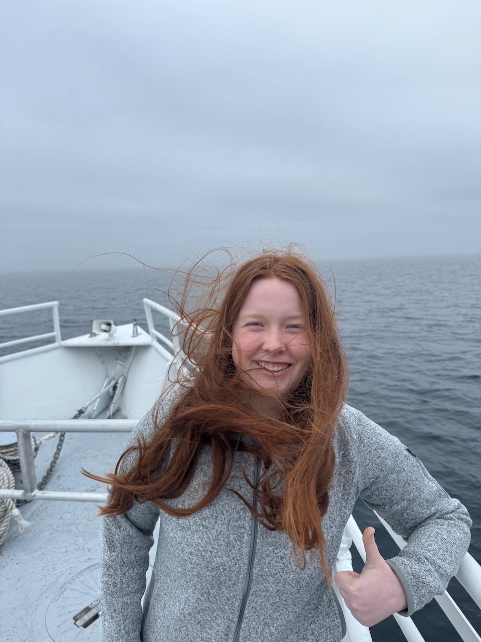 Cameron with her hair blowing in the wind, giving a big thumbs up, standing outside on the deck of the Queen IV as we make our way to the island.