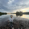 Cameron standing outside in hiking cloths, at Moose Point in Isle Royale at sunset. The sky is alive with color that is reflected into Tobin Harbor behind us.