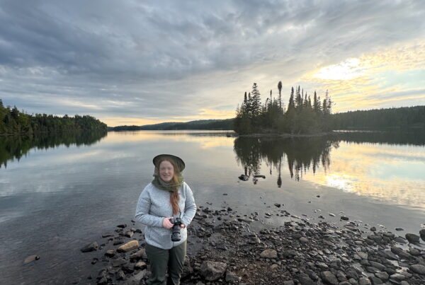 Cameron standing outside in hiking cloths, at Moose Point in Isle Royale at sunset. The sky is alive with color that is reflected into Tobin Harbor behind us.