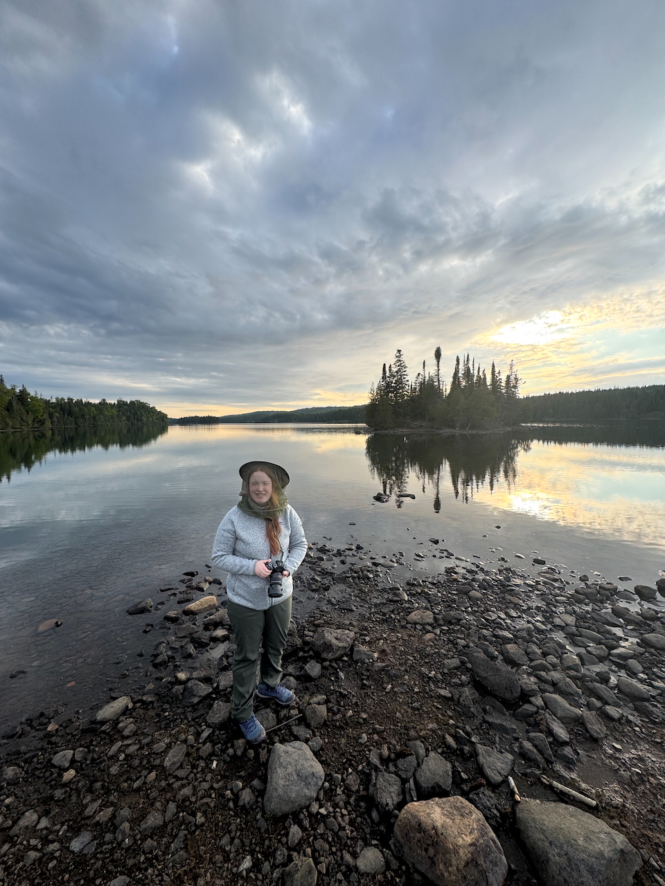 Cameron standing outside in hiking cloths, at Moose Point in Isle Royale at sunset. The sky is alive with color that is reflected into Tobin Harbor behind us.