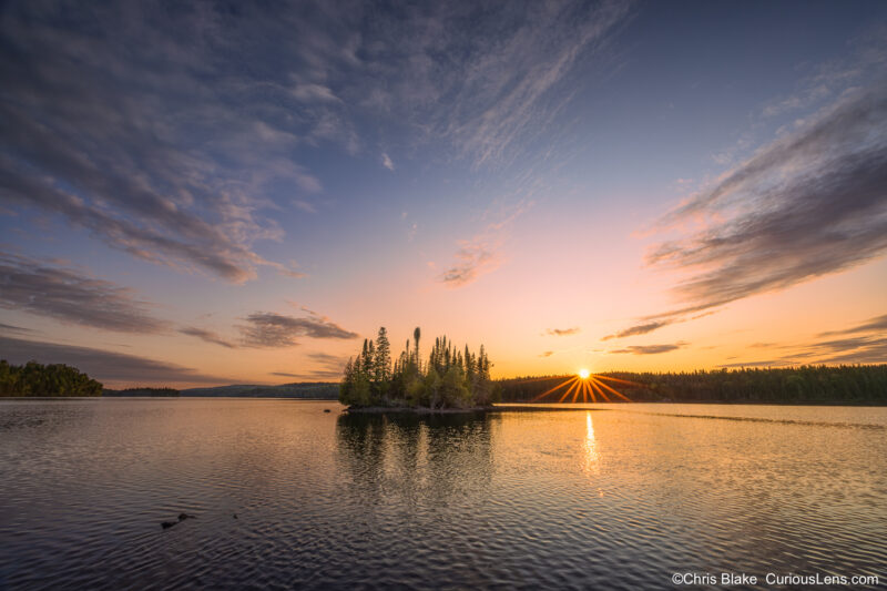 Sunset over Tobin Harbor in Isle Royale National Park with a small forested island in the foreground and a sunburst just above the tree line