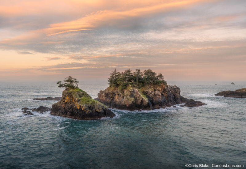 Panoramic sunrise photo at Seal Cove, Oregon, showing two massive sea stacks with trees and glowing morning clouds over the Pacific.