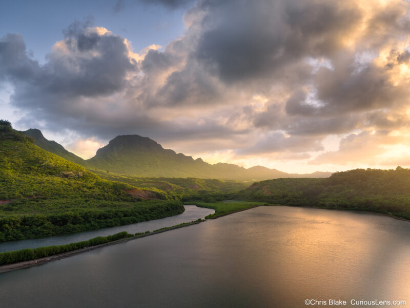 Sunset over Alekoko Fishpond in Kauai with storm clouds and golden light breaking through onto the mountains and trees.
