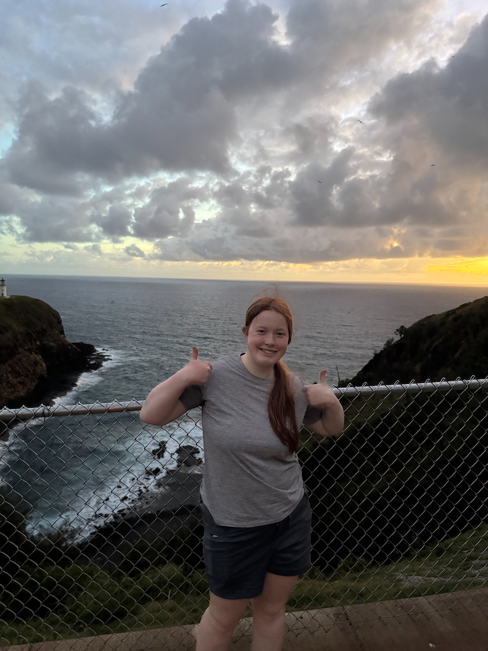 Cameron standing at Kīlauea Lighthouse at sunrise with ocean cliffs behind her.