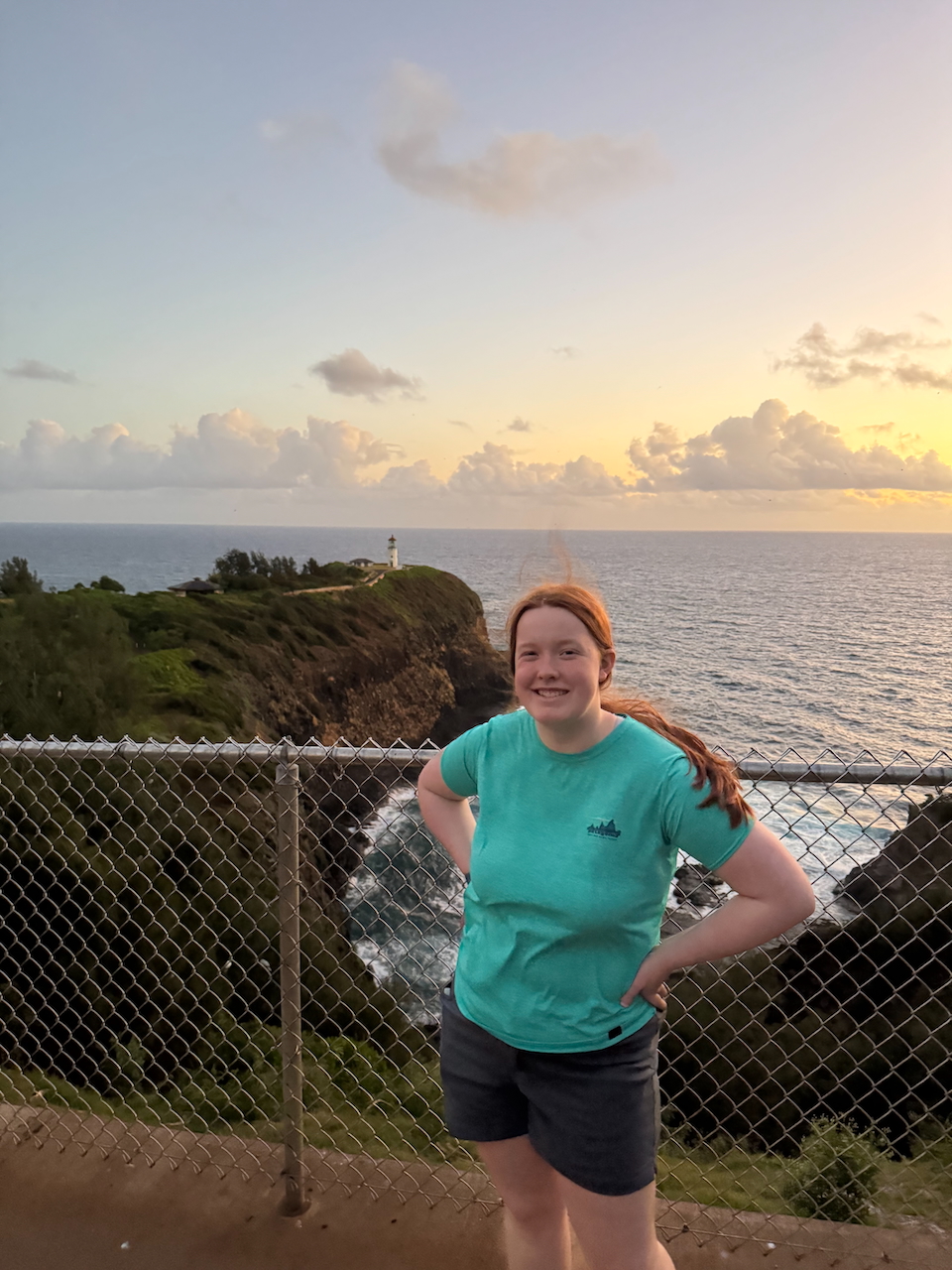 Cameron smiling at sunrise with the Kīlauea Point Lighthouse and coastal cliffs in the background.