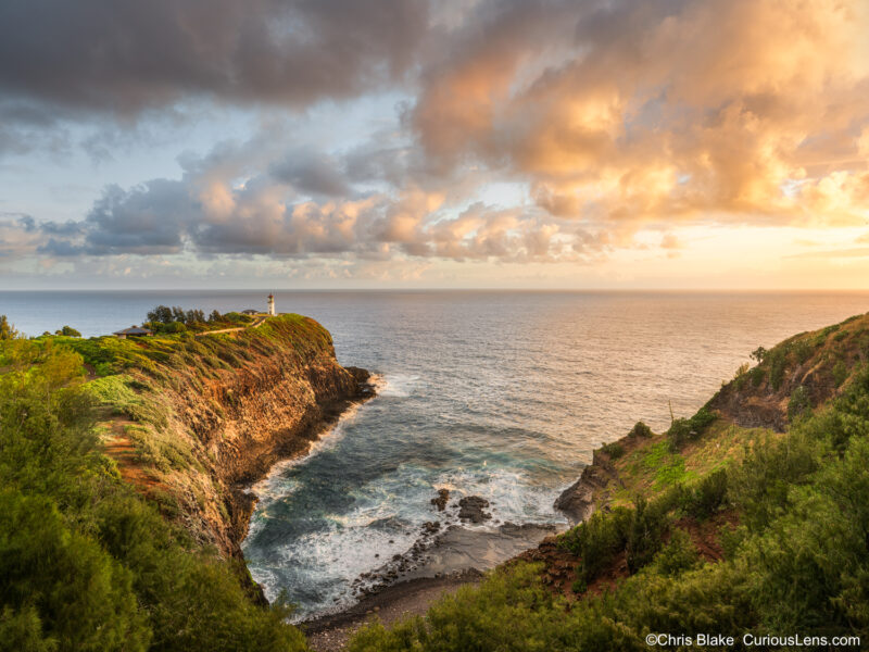 Wide sunrise view of Kilauea Lighthouse cove on Kauai with storm clouds, glowing light on cliffs, and seabirds along the coastline.