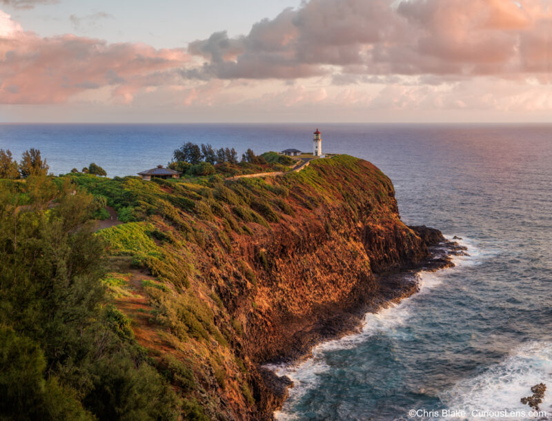 Panoramic sunrise of Kilauea Lighthouse on Kauai with warm morning light glowing on cliffs, ocean waves, and lighthouse against soft sky.