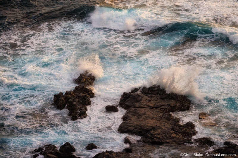 Crashing surf on black volcanic rocks below Kilauea Point, with ocean spray glowing in sunrise light.