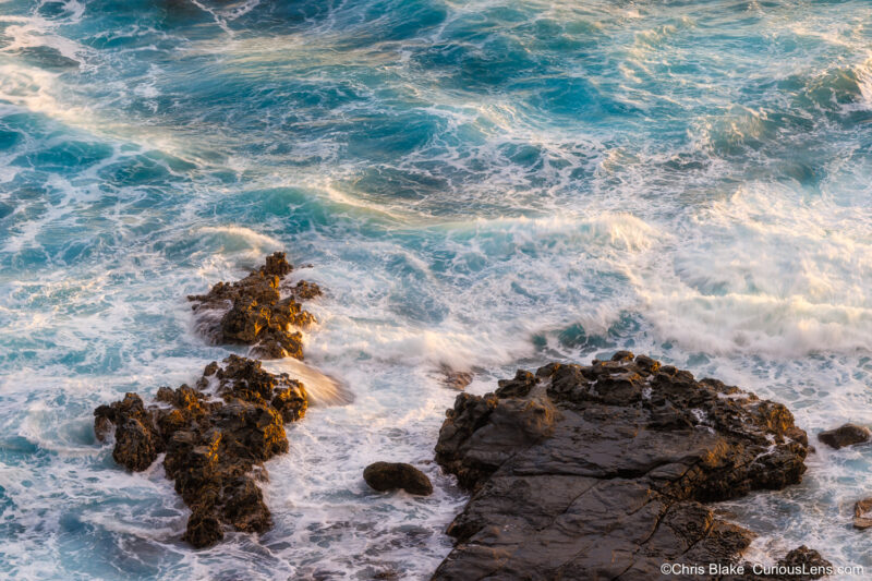 Turquoise sunrise surf surging over black lava rocks below Kilauea Point on Kauai, with golden light highlighting the rugged coastline.