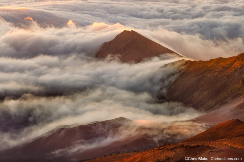 Sunrise over Haleakalā Crater on Maui, with morning light striking the clouds and red volcanic rock.