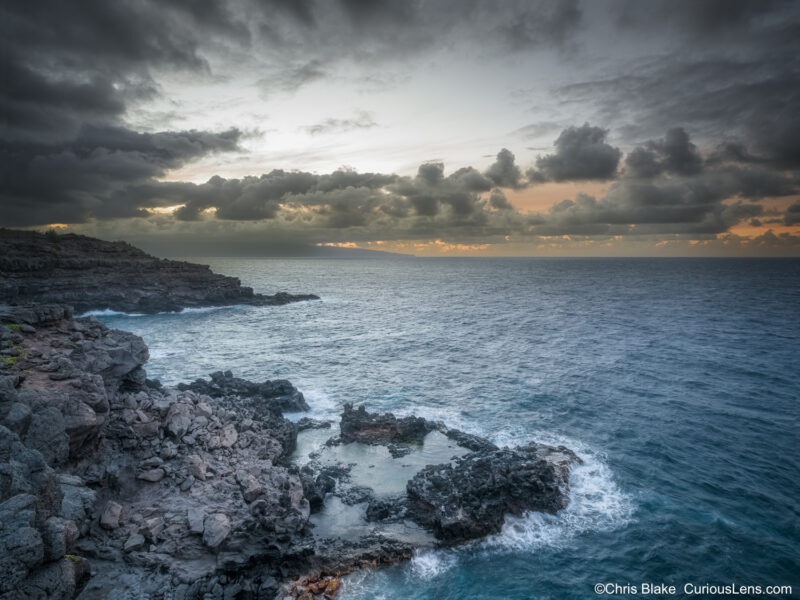 Storm clouds over the rocky coastline near Nakalele Blowhole on Maui, with waves crashing into tidepools at dusk.