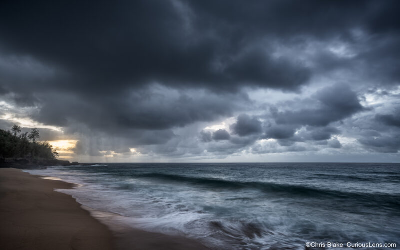 Stormy twilight sky over Secret Beach on Kauai with waves rolling onto the sand and rain falling at sunset.