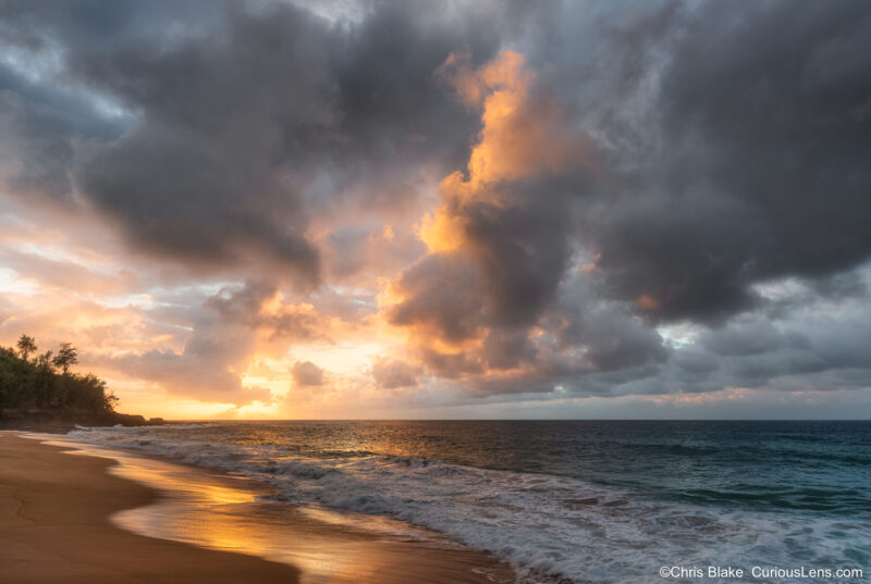 Storm clouds breaking at sunset over Secret Beach on Kauai, with waves retreating across wet sand reflecting golden light.