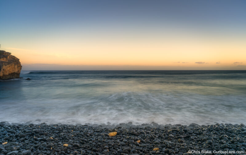 Blue hour at Shipwreck Beach on Kauai with smooth ocean waves, calm horizon, and black pebbles with scattered yellow stones on the shore.