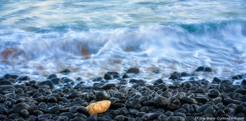 Wave washing over black lava stones at Shipwreck Beach on Kauai, with one golden rock standing out in contrast.