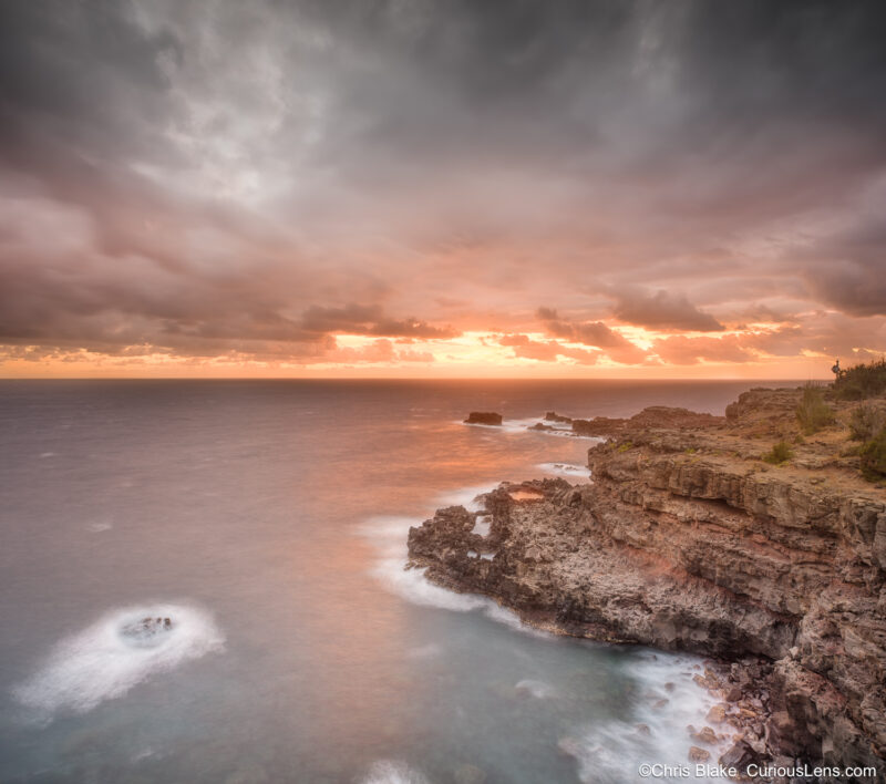 Sunrise over the Pacific Ocean at Nakalele Blowhole on Maui, with dramatic clouds and red reflections on the water.