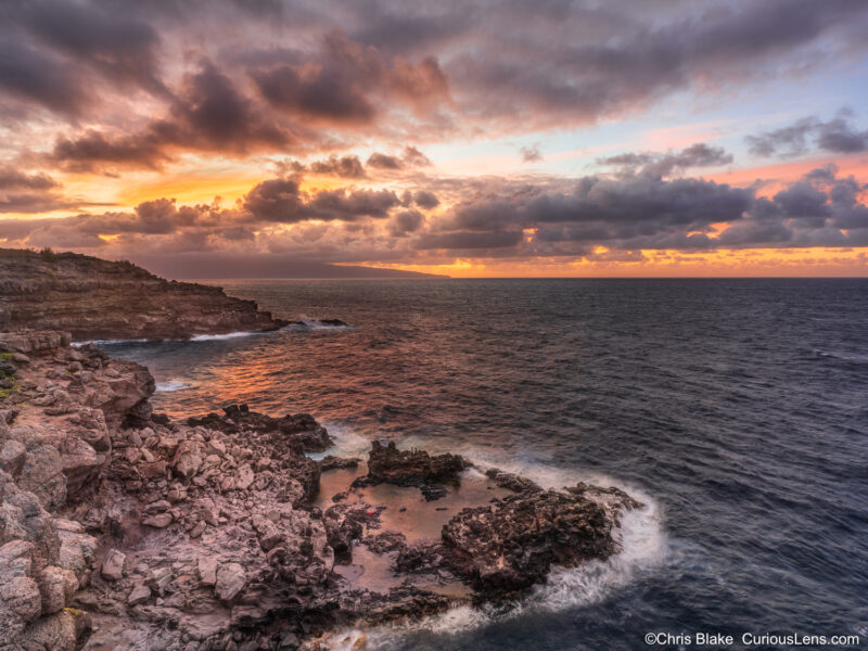 Fiery sunset over the cliffs near Nakalele Blowhole on Maui, with golden-red light reflecting on clouds and ocean.