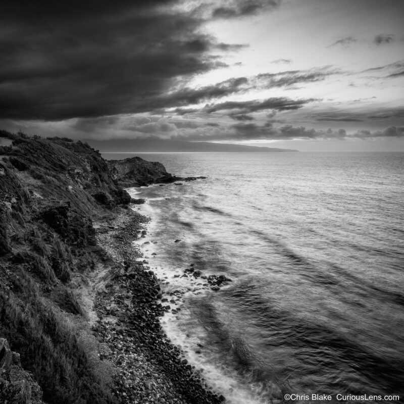 Black and white photograph of the Lipoa Ridge coastline on Maui at sunset, with storm clouds overhead and waves moving against the rocky shore.