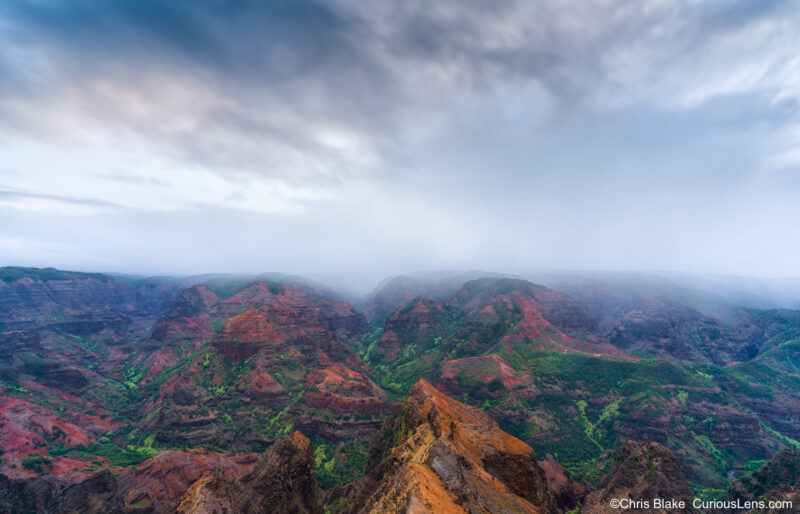 Clouds rolling over Waimea Canyon at sunrise, with red cliffs and green slopes visible beneath the mist.