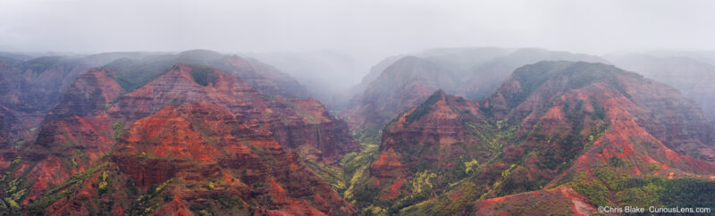 Panoramic view of Waimea Canyon at sunrise, with red cliffs and green slopes fading into mist.