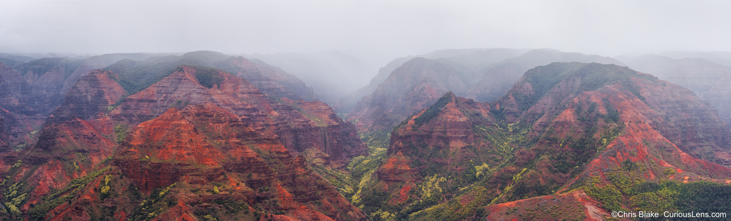 Panoramic view of Waimea Canyon at sunrise, with red cliffs and green slopes fading into mist.