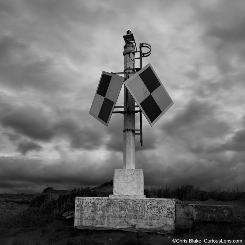 Weathered lighthouse marker at Nakalele Blowhole on Maui, standing against dark storm clouds.