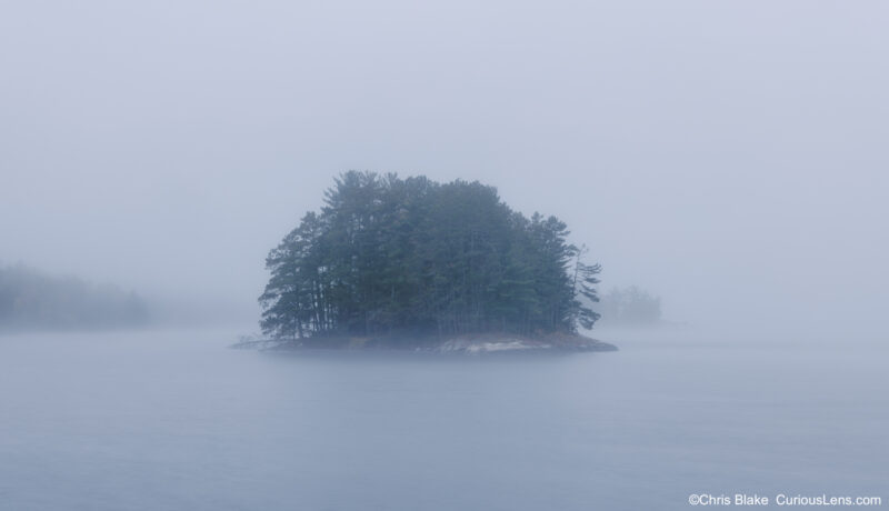 A small pine-covered island on Kabetogama Lake emerges from thick morning fog at sunrise in Voyageurs National Park, Minnesota.